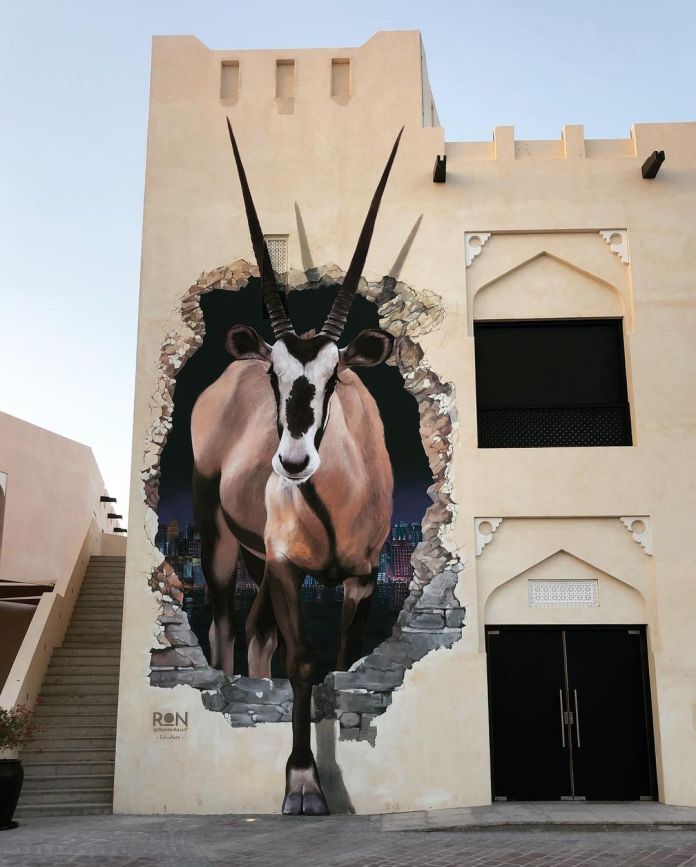 A striking mural titled "Oryx Going Ahead" by Martín Ron, located in Doha, Qatar. The artwork depicts a majestic oryx, a symbol of Qatar, appearing to step through a crumbling wall, blending hyper-realistic art with architectural features. The mural creates an illusion of depth, with the oryx emerging from a nighttime cityscape visible through the hole in the wall. The beige, traditional-style building contrasts sharply with the vivid details of the oryx and the urban backdrop, highlighting the seamless integration of art into its environment. The bold design embodies movement and cultural significance.