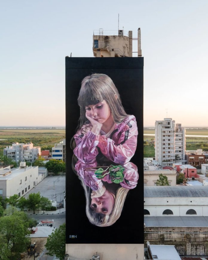 A large mural titled "Reflections" by Martín Ron, located in San Nicolás de los Arroyos, Argentina. The artwork features a young girl in a pink jacket, resting her head on her hand in a pensive pose. Her image is mirrored below, creating a symmetrical and reflective effect, with the lower reflection holding a small green plant. The mural is painted on the side of a tall building, standing out against the surrounding cityscape and the expansive green and golden fields in the distance. The hyper-realistic details and emotional depth of the piece draw attention to its urban environment.