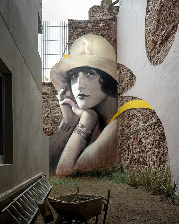 A nostalgic mural by Martín Ron in San Telmo, Buenos Aires, Argentina, featuring a striking portrait of a woman in a vintage style. She wears a soft yellow hat and bracelets, her pose exuding elegance as her face rests gently on her hand. The artwork is seamlessly integrated into a partially exposed brick wall, blending old-world charm with modern urban aesthetics. Yellow accents add a touch of vibrancy to the muted tones. The mural is located in a narrow courtyard surrounded by buildings, with construction tools and a wheelbarrow in the foreground, further enhancing the mural's connection to its environment.