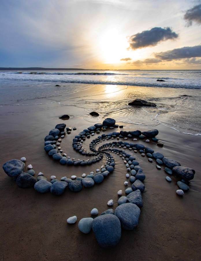 Land artist Jon Foreman sitting beside a large stone spiral on a beach in Druidston, Wales, with black stones arranged in concentric rings that decrease in size toward the center.