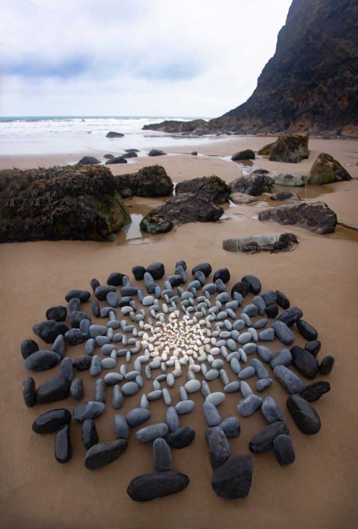 Circular stone artwork on a beach featuring a sunburst design with white pebbles in the center and darker stones radiating outward, surrounded by rocky shoreline and waves in the background.
