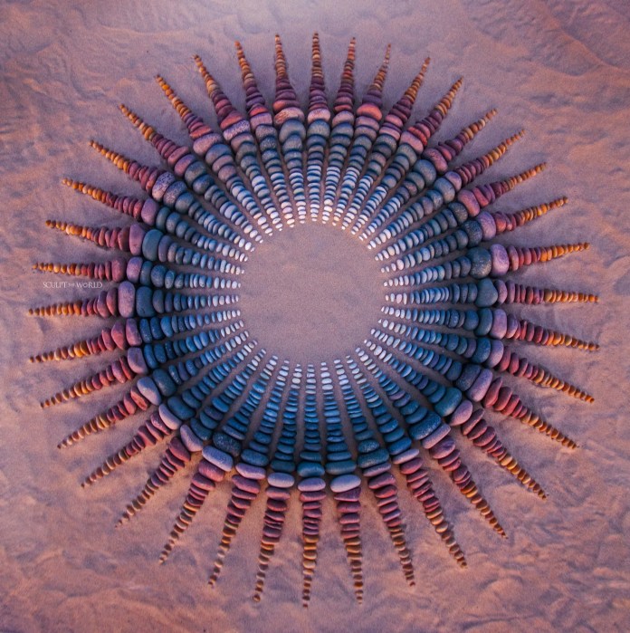 Color gradient pebble circle on sand, shifting from white and gold in the center to orange, red, purple, and blue toward the edges in symmetrical layers.