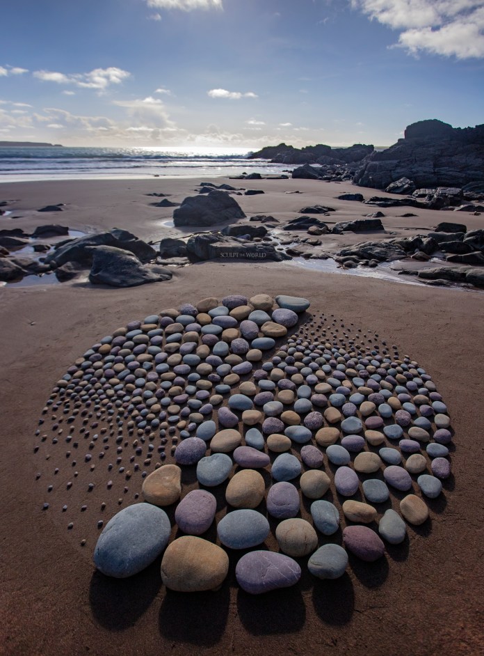 Stone sculpture on a Welsh beach showing a circular form visually halved with mirrored sides of blue-grey and tan pebbles under a bright sky.