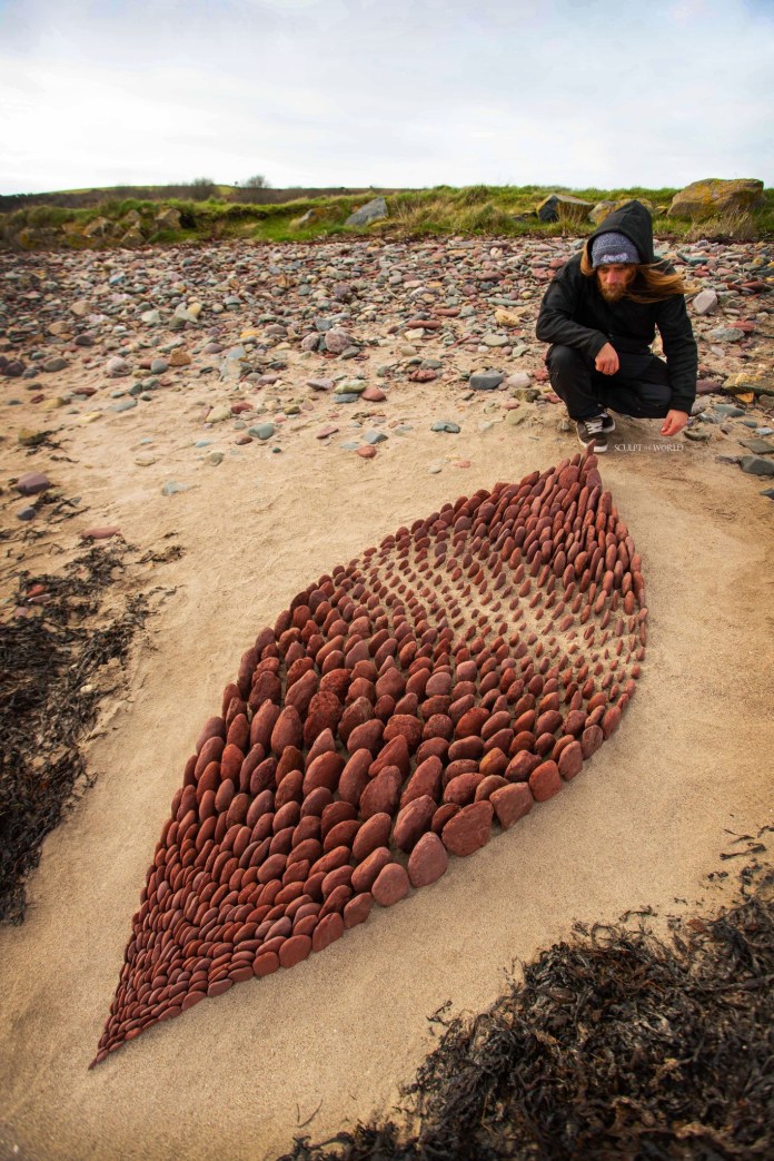 Leaf-shaped land art made of reddish stones in gradually changing sizes, arranged in rows on a sandy beach near scattered pebbles and seaweed.