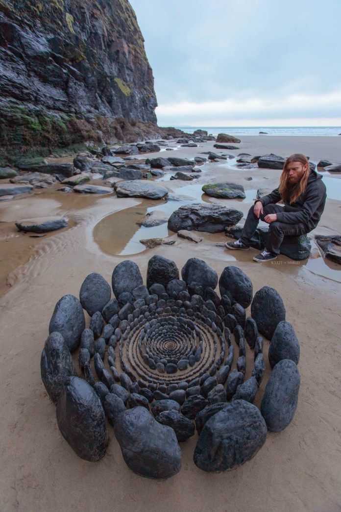 Expansive stone arrangement on a Welsh beach with concentric rings transitioning from white in the center to black stones along the outer edges.