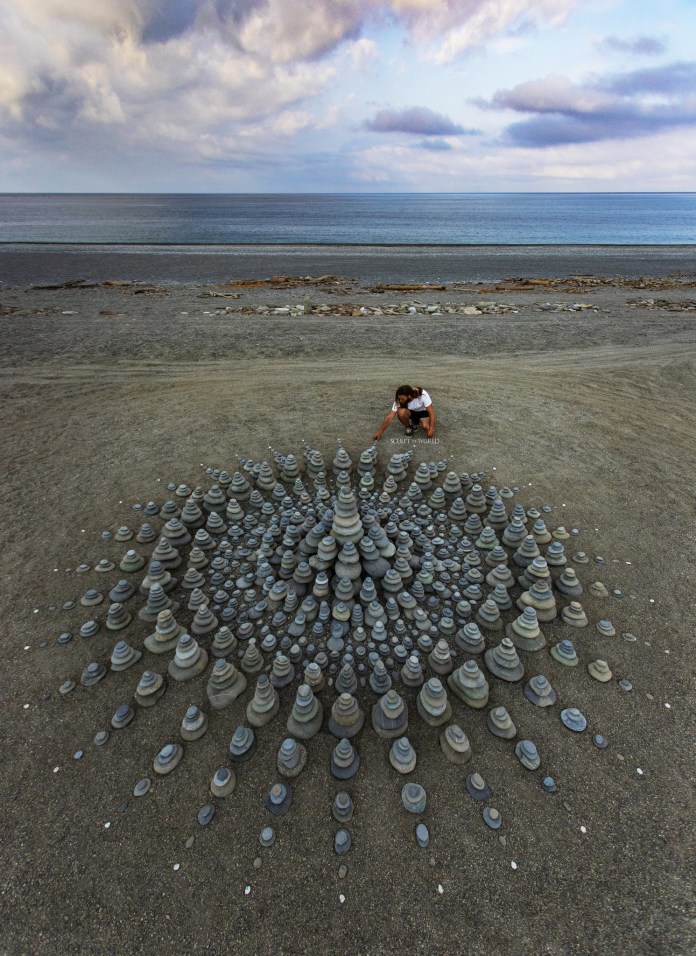 Spiral stone artwork at the water’s edge, made of alternating dark and white stones forming twisting arms with ocean waves and a glowing horizon behind.