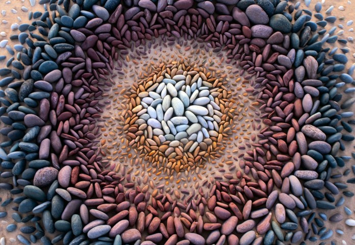 Dozens of tiny balanced stone stacks forming a symmetrical radial pattern on a pebble-covered beach, with an artist kneeling beside it.