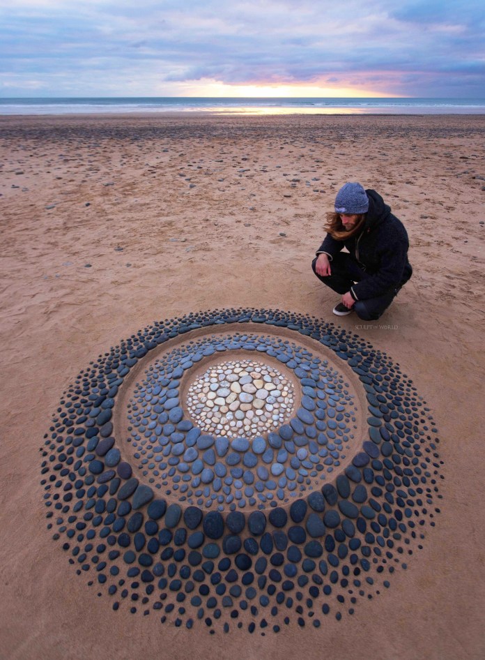 Stone mandala in a sunburst layout with colorful rays extending outward from a hollow center, arranged on smooth sand under soft sunset light.