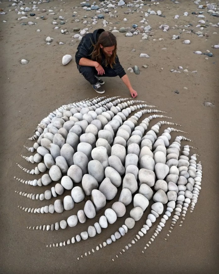 Jon Foreman crouching beside his beach artwork Fluidform at Pensarn, Wales—featuring rows of white stones increasing and decreasing in size to create a fluid, radial shape that seems to flow outward across the wet sand.