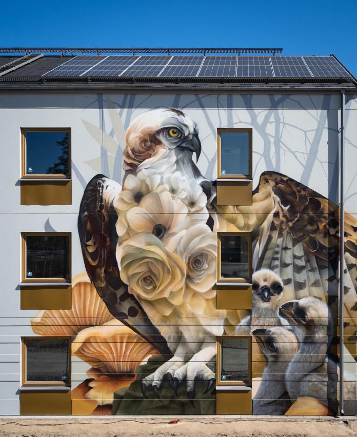 Mural of an osprey made from white roses and shells with two chicks below, painted across a building facade in Nykvarn, Sweden.