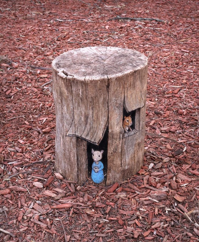 Chalk and paint artwork on a tree stump depicting two mice peeking out from carved openings, one in a blue dress holding a teacup and one above in shadow, surrounded by red mulch.