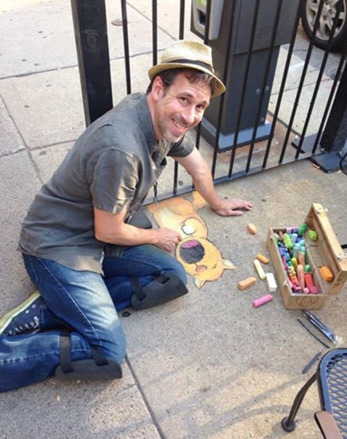 David Zinn, a street artist known for his whimsical chalk art, is captured mid-creation on a city sidewalk. He is kneeling beside a partially completed chalk drawing of a cartoon-style dog, vibrant with colors like yellow and pink. Zinn is dressed casually in a gray shirt, jeans, and a straw hat, with a box of multicolored chalks open next to him. The artwork is situated near a black metal fence and a parking meter, showcasing his knack for blending street elements with his art.