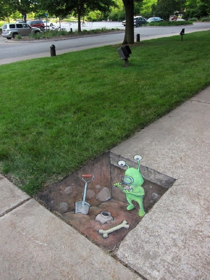 A clever chalk art piece by David Zinn transforms a section of a sidewalk into an excavation site featuring a green alien with antennae. The alien is depicted curiously examining a bone while standing inside a hole that appears to be dug into the pavement. Beside the alien, a red-handled shovel and an unearthed artifact add to the illusion. The artwork is bordered by grass, a tree, and a parking lot in the background, perfectly integrating Zinn's imaginative creation with the urban landscape.