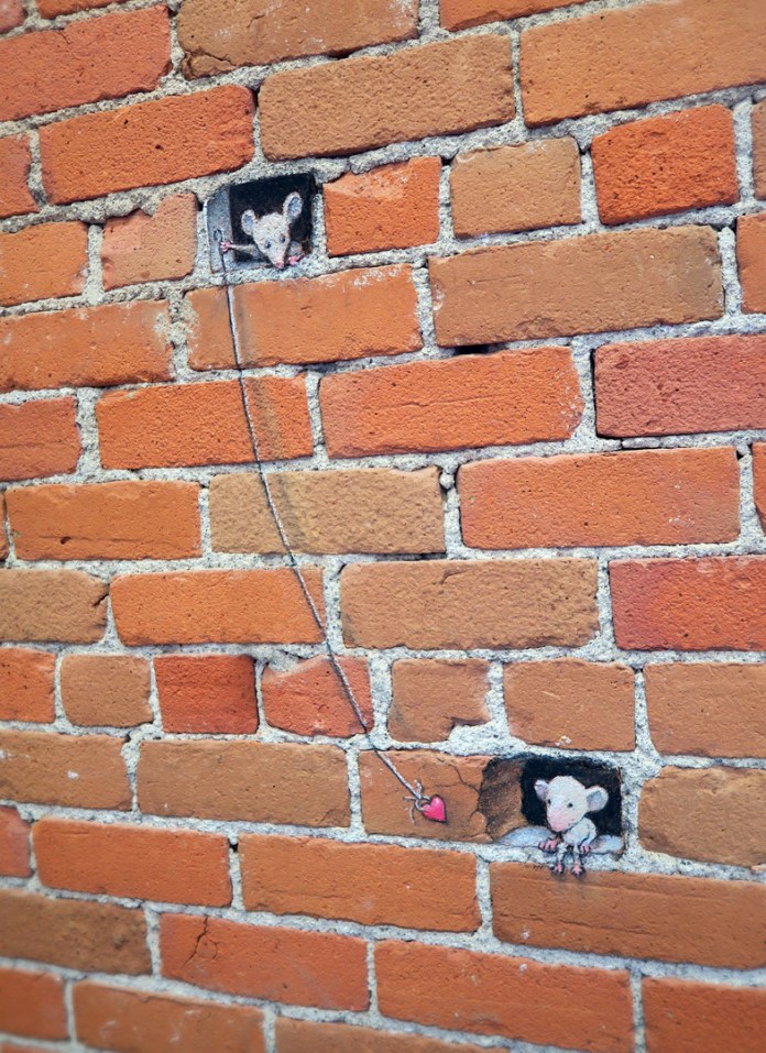 A creative chalk art piece by David Zinn incorporates a red brick wall, transforming it into a tiny scene of interaction. Two mice are drawn peeking out of rectangular gaps in the wall, connected by a piece of string holding a dangling red heart. One mouse reaches toward the heart, while the other observes from above, adding a touch of emotion and whimsy. The vibrant red bricks and textured mortar provide a striking background, highlighting Zinn's ability to blend his art seamlessly into urban environments. The scene conveys a sense of connection and curiosity in a playful, imaginative manner