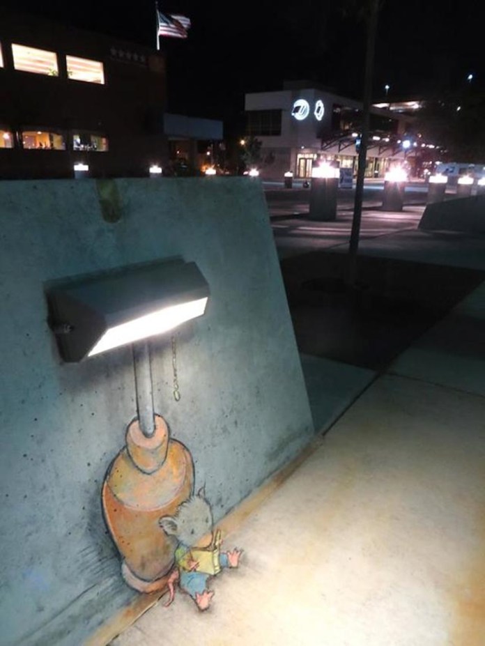 A heartwarming chalk art piece by David Zinn, featuring a small mouse sitting under an outdoor wall light at night. The mouse, drawn with soft fur and pink paws, is leaning against the light's base while reading a book, illuminated by the glow. The light fixture, seamlessly incorporated into the art, resembles a cozy lamp. The nighttime setting includes a background of lit buildings and an American flag waving in the distance, adding depth and atmosphere to the scene. Zinn’s work evokes a sense of warmth and quiet storytelling.