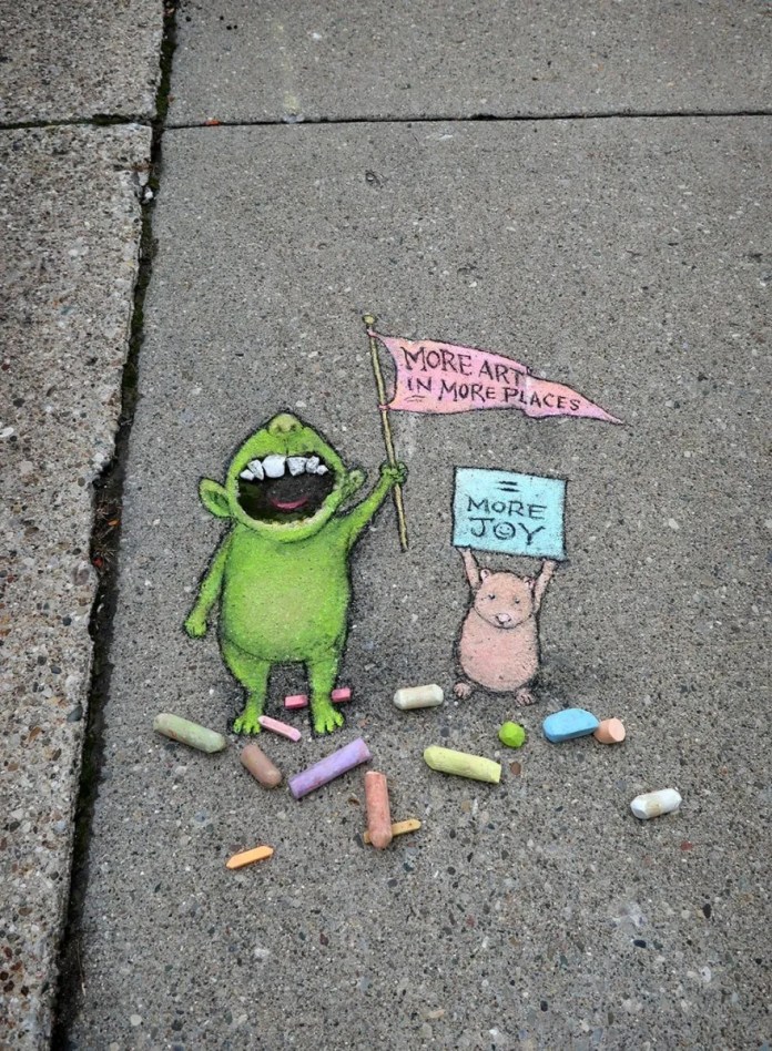 A cheerful chalk art scene by David Zinn featuring a green monster and a small pink mouse on a sidewalk. The green monster is joyfully waving a pink flag that reads, "More Art in More Places," while the mouse holds a blue sign that says, "= More Joy." The artwork is surrounded by scattered colorful chalk pieces, adding a playful and spontaneous vibe. The rough texture of the sidewalk enhances the handmade charm of the piece, delivering an uplifting message about the power of art to bring happiness.