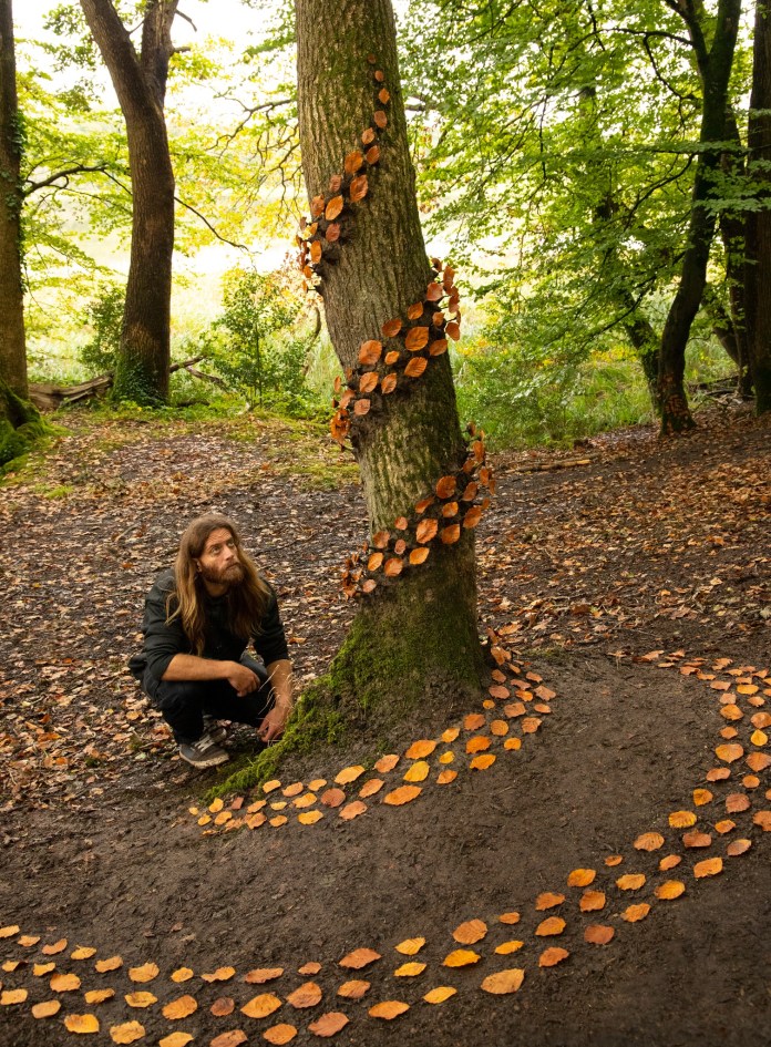 A golden spiral made of small yellow leaves arranged on a dirt clearing, surrounded by fallen brown leaves in a wooded area. The artist crouches nearby observing the work.