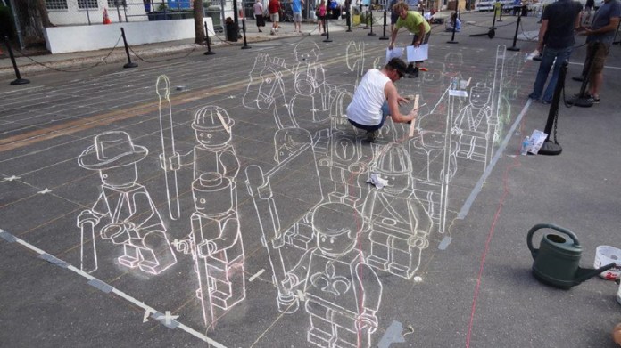 3D chalk art by artists Leon Keer, Ruben Poncia, Remko van Schaik, and Peter Westerink at the 4th Sarasota Chalk Festival in Florida, USA. The artwork features a group of LEGO-like characters drawn on the pavement, appearing to stand in formation with weapons and armor, resembling a medieval or fantasy army. The outlines and shading create a layered effect that gives depth and dimension to each figure. One of the artists is seen in the process of drawing, adding intricate details to bring the illusion to life, while spectators observe the work in progress.