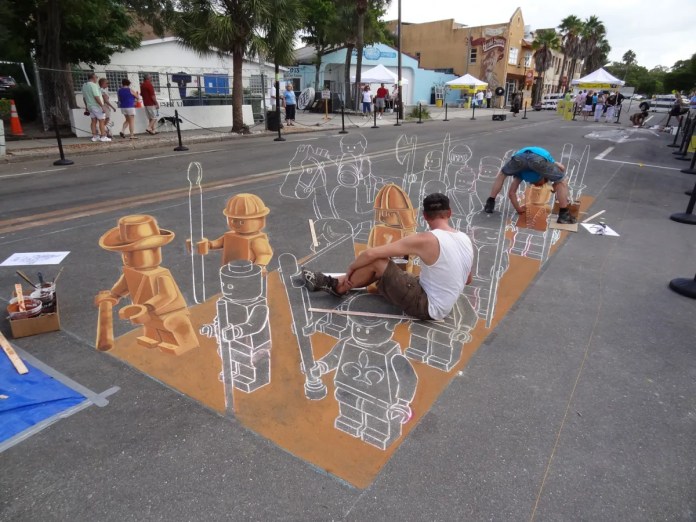 Artists Leon Keer, Ruben Poncia, Remko van Schaik, and Peter Westerink working on a 3D chalk mural at the 4th Sarasota Chalk Festival in Florida, USA. The artwork depicts a procession of LEGO figures dressed as knights, soldiers, and adventurers, some already colored in shades of brown and gold, giving the illusion of three-dimensional plastic figures standing on the street. Two artists are seated within the artwork, meticulously adding color and detail to each character, enhancing the sense of depth and realism, while onlookers observe the creative process.