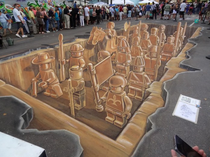 Finished 3D chalk art by artists Leon Keer, Ruben Poncia, Remko van Schaik, and Peter Westerink at the 4th Sarasota Chalk Festival in Florida, USA. The artwork depicts an army of LEGO figures, including knights, soldiers, and adventurers, standing in a sunken pit drawn onto the street. The illusion of depth and shading makes it appear as though the characters are within a carved-out trench, with detailed textures on each figure. Spectators gather around to view the immersive piece, which captivates with its impressive realism and playful use of childhood imagery in a large-scale street mural.