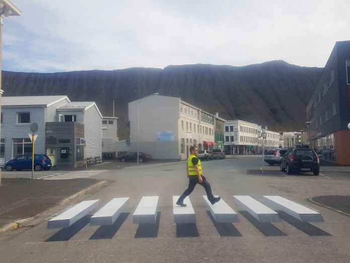 3D pedestrian crossing in Iceland designed to reduce speeding by creating an optical illusion of floating white blocks. Painted on the street, the crosswalk appears three-dimensional, with each white stripe looking like it hovers above the ground. A man in a yellow safety vest walks across, adding to the illusion's effectiveness and highlighting the creative approach to traffic safety. The surrounding street is framed by small buildings and a mountainous backdrop, emphasizing the quiet, small-town setting of this innovative design.