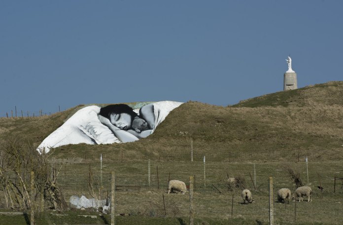Land art mural titled 'The Sleeping Beauty' by Made in Graffiti, located on a hillside in Picardie, France. The artwork depicts a serene black-and-white portrait of a woman sleeping, nestled against the natural contours of the hill, blending art with the landscape. In the distance, a statue stands on a concrete structure, adding a sense of scale and depth. Sheep graze in the foreground, enhancing the peaceful rural setting. The mural’s simplicity and integration into the environment evoke a sense of tranquility and harmony with nature.