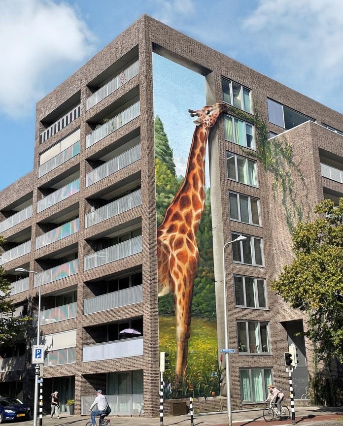 Mural titled 'Giraffe Eating the Plants' in Utrecht, Netherlands, depicting a realistic giraffe reaching up to nibble on plants growing on the balcony of a residential building. The artwork covers the corner of the building, creating an optical illusion that integrates the giraffe seamlessly with the architectural structure. The background of the mural shows lush greenery, blending urban and natural elements in a playful way. Cyclists and pedestrians in the foreground add to the urban setting, while the towering giraffe mural adds whimsy and charm to the neighborhood.
