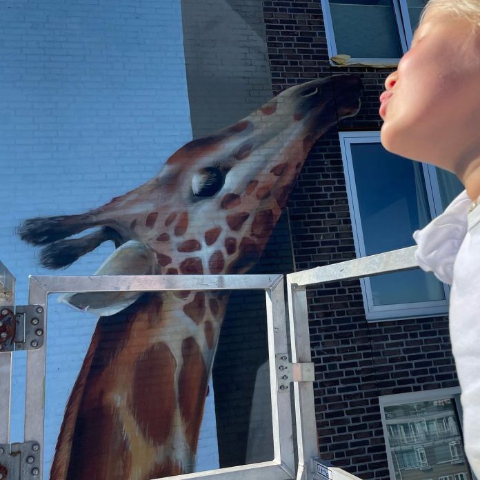 A whimsical perspective photo of Jan Is De Man's giraffe mural in Utrecht, Netherlands, showing a close-up of the giraffe's head reaching toward greenery, with a child in the foreground playfully mimicking feeding the giraffe, adding a playful and interactive element to the artwork.