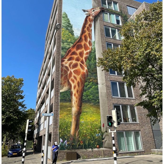 A side-angle shot of Jan Is De Man's mural in Utrecht, Netherlands, where a giraffe painted on the wall appears to feed on green plants creeping up the adjacent wall, blending art with nature.