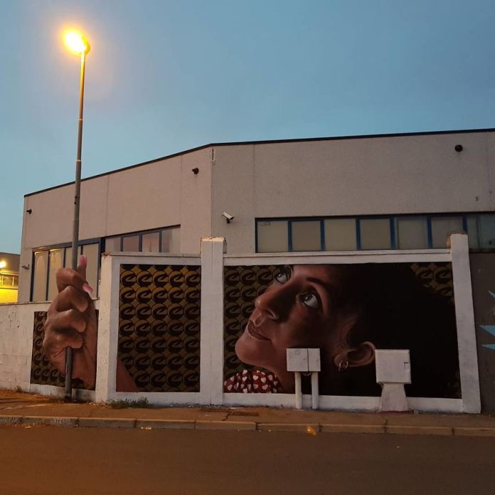 A striking mural by Cosimo Cheone Caiffa, featuring a hyper-realistic portrait of a woman wearing a red polka-dot blouse. The woman gazes thoughtfully upward at a lamppost that appears to be part of the artwork, seamlessly integrating the real-world structure into the painting. Her hand is depicted as holding the lamppost, enhancing the illusion. The mural's detailed shading and lifelike features, combined with a patterned golden backdrop, create a visually stunning and immersive scene, transforming the urban setting into a captivating work of art.