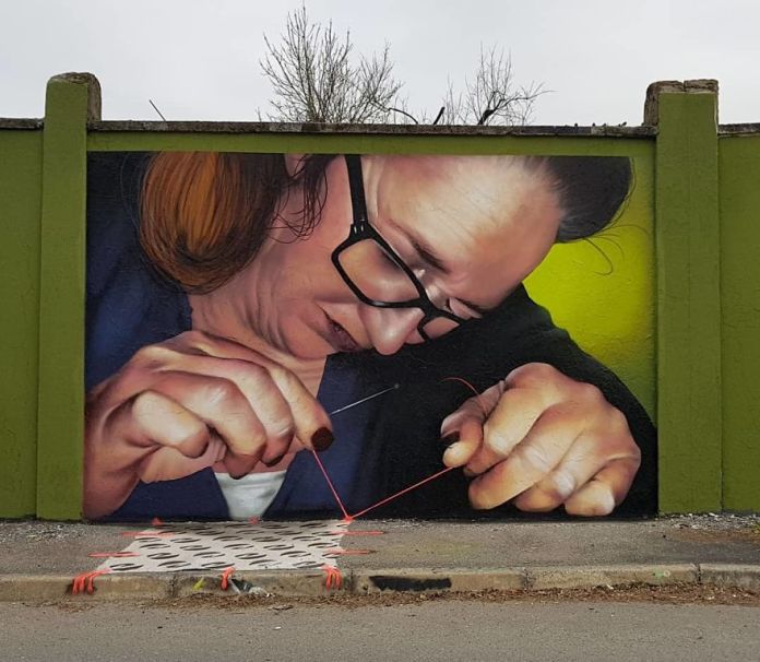 A detailed 3D mural by Cosimo Cheone Caiffa depicting a woman sewing, with a red thread extending from her needle onto the ground. The hyper-realistic artwork shows the woman wearing glasses, deeply focused as she pulls the thread through a needle, creating an intricate design that flows seamlessly into the real-world pavement. The green background contrasts with the lifelike portrayal, emphasizing her hands and the fine details of the thread. This clever integration of painted and physical elements transforms the space into an engaging and immersive piece of street art.