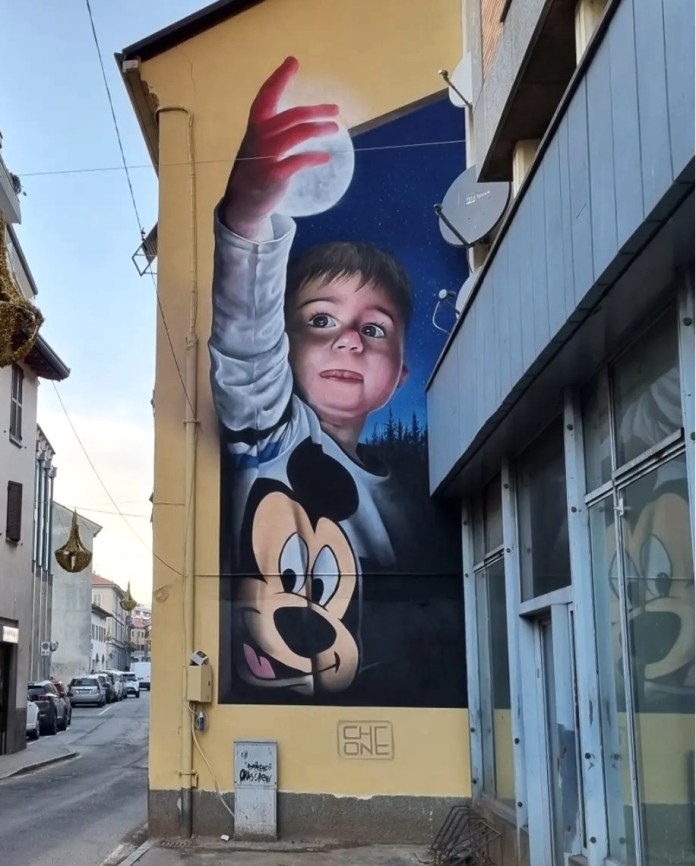 A whimsical 3D mural by Cosimo Cheone Caiffa depicting a young boy reaching out to touch a glowing moon, set against a starry night sky. The boy's wide-eyed expression and outstretched hand convey a sense of wonder and curiosity, while a playful depiction of Mickey Mouse is featured prominently on his shirt. The combination of realism in the boy's features and the fantastical elements of the moon and night scene creates an enchanting and dreamlike composition. The mural transforms the urban wall into a heartwarming and magical piece of art.