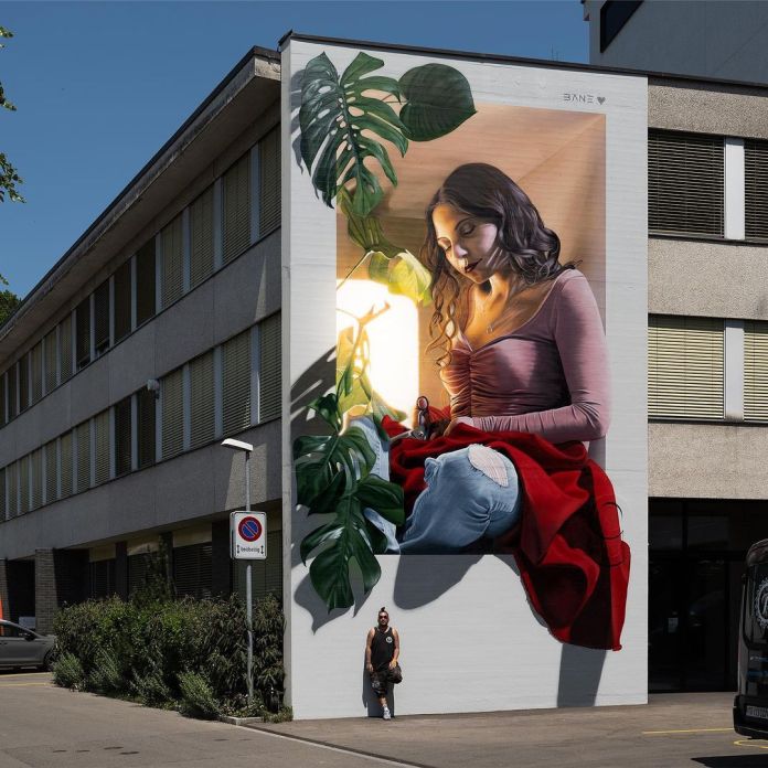 An awe-inspiring 3D anamorphic mural titled 'Cut Through History' by Fabian Bane Florin in Frauenfeld, Switzerland. The artwork depicts a serene woman sitting with a thoughtful expression, surrounded by large green monstera leaves. She holds a piece of fabric and sewing tools, symbolizing the act of stitching history together. Her red shawl and ripped jeans add texture and realism, while warm light bathes the scene, enhancing its depth. Painted on the side of a modern building for the Street Art Festival Frauenfeld, the mural captivates with its intricate details and profound narrative.