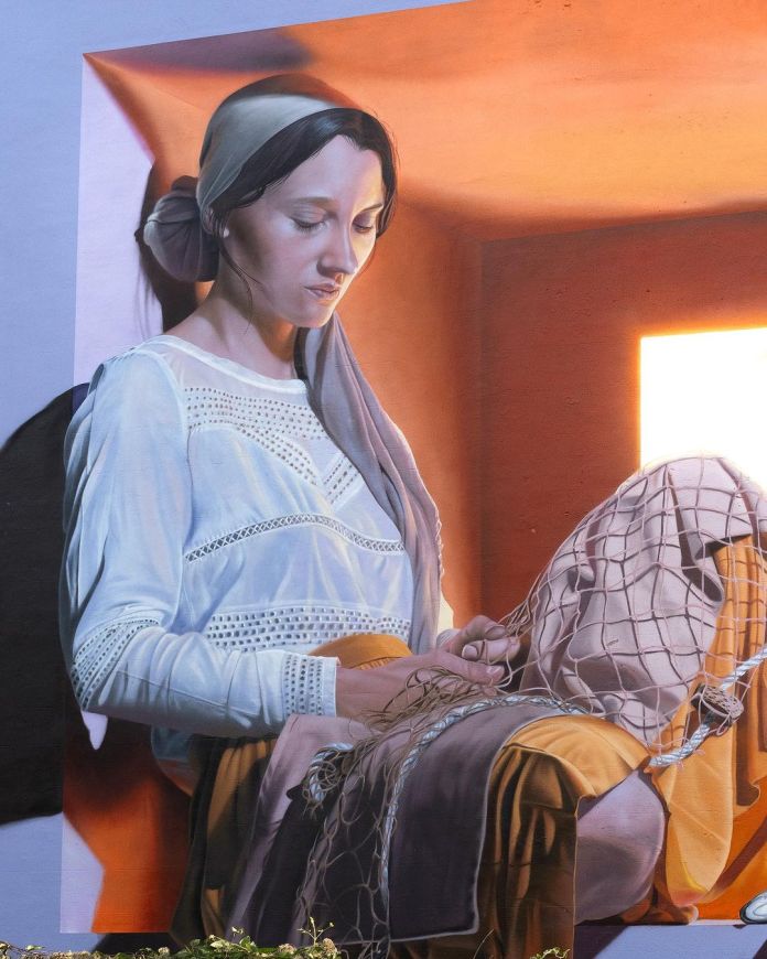 A detailed close-up of 'The Fisher Girl' mural by Fabian Bane Florin in Mons, Belgium. The portrait focuses on the young woman’s serene face and hands as she carefully mends a fishing net. Her delicate lace blouse and headscarf are rendered with remarkable realism, highlighting her peaceful expression. The sunlight streaming through the window softly illuminates her features, emphasizing the intricate details of the artwork and the warm, contemplative mood it conveys.