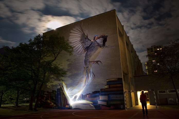 A stunning nighttime view of the mural 'I Have a Dream' by Fabian Bane Florin and Pest in Chur, Switzerland. Illuminated by soft light, the artwork features an open book with its pages transforming into a soaring sparrow carrying a young girl. The glowing effect of the book contrasts beautifully with the surrounding darkness, emphasizing the mural's dreamlike quality. The scene is framed by trees under a cloudy night sky, enhancing the magical atmosphere of the piece. A person stands nearby, gazing at the mural, adding scale and a sense of wonder.