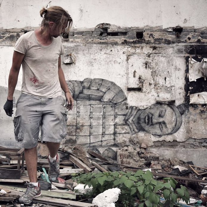 Seth Globepainter stands amidst rubble in front of a partially crumbled wall featuring his mural of a fallen terracotta warrior. The mural merges seamlessly with the raw, textured surface of the wall, blending historical themes with urban decay. Seth, dressed casually in paint-splattered clothes, looks at his creation, which contrasts with the surrounding debris and plants growing through the wreckage.