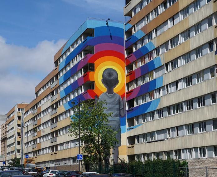 A vibrant mural by Seth Globepainter titled "Enter the Vortex," located on rue Jeanne d’Arc in Paris 13, France, created in collaboration with Galerie Itinerrance. The artwork features a boy seen from behind, staring into a hypnotic rainbow spiral that spans across a multi-story building, blending bold colors with the structure’s geometry. The piece transforms the urban environment, drawing attention to both the artwork and its surrounding space.