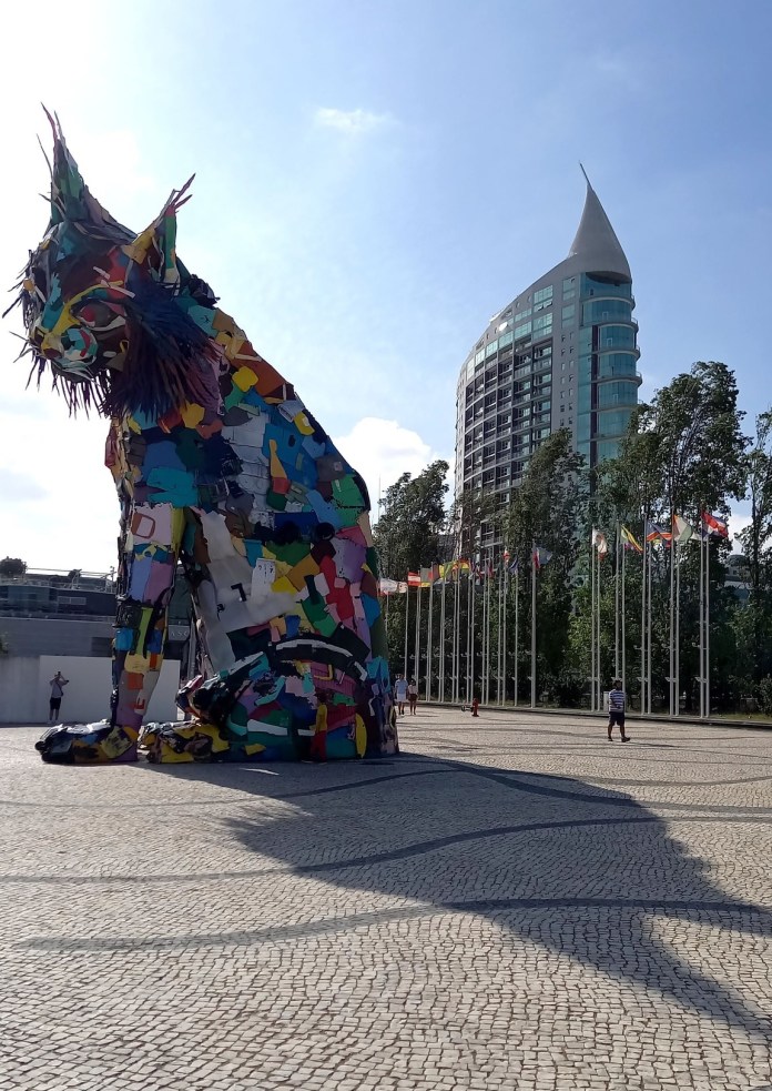 A towering sculpture of a lynx made entirely from colorful recycled plastic waste, created by the artist Bordalo II, stands in Lisbon, Portugal. The artwork, rich with vibrant hues, demonstrates the fusion of art and environmental awareness. The sculpture is located in an open plaza with cobblestone paving, with a modern curved building and flagpoles in the background, emphasizing the urban setting.