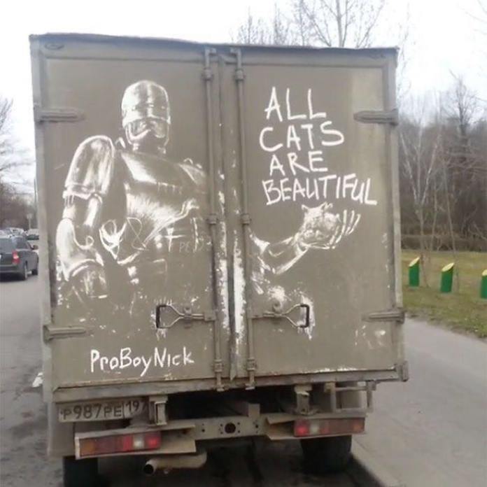 Creative grime art on the back of a truck by artist ProBoyNick. The artwork features a RoboCop-like figure holding a small cat, alongside the playful phrase 'ALL CATS ARE BEAUTIFUL,' etched into the dirt on the truck door. ProBoyNick’s signature style combines pop culture references with humor, transforming dirty vehicle surfaces into canvases.