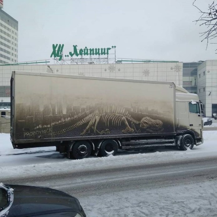 Grime art by ProBoyNick on the side of a truck, depicting a dinosaur skeleton stretched across the length of the trailer. The detailed artwork uses the dirt on the truck’s surface to create a fossil-like effect, set against a faint cityscape background. The truck is parked in a snowy urban environment, transforming its exterior into a striking piece of mobile art.