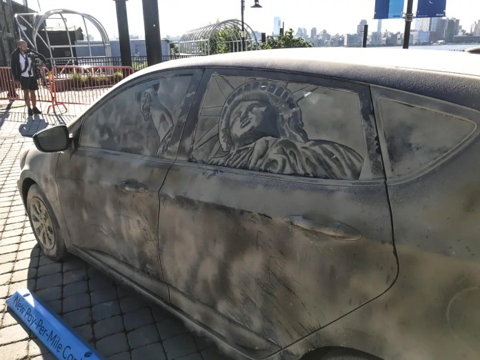 Grime art on the side windows of a dusty car, featuring an intricately etched image of the Statue of Liberty. The iconic figure is carefully crafted into the dirt on the windows, creating a striking and patriotic display. The car is parked on a cobblestone surface with a city skyline visible in the background, showcasing how grime art transforms ordinary vehicles into moving canvases.