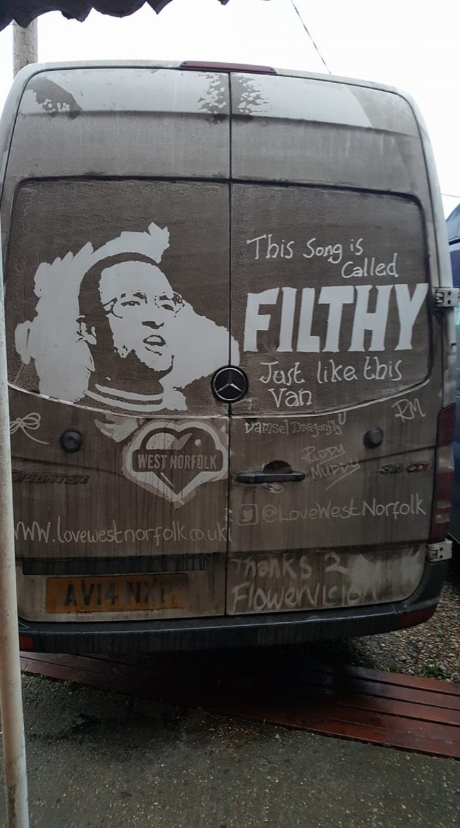 Grime art on the back of a van, featuring a portrait and the humorous phrase, 'This Song is Called FILTHY, Just like this Van.' The artwork includes a logo for 'West Norfolk' and several tags, including social media handles like '@LoveWestNorfolk' and phrases like 'Thanks 2 Flowervision.' The artist cleverly incorporates the van’s dirt to convey a playful message, turning the vehicle into a unique canvas.