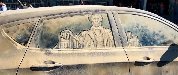 Grime art on the side windows of a dusty car, depicting Abraham Lincoln seated as he appears in the Lincoln Memorial. The detailed portrait is etched into the dirt, capturing Lincoln's likeness with his hands resting on the arms of a grand chair. The artwork transforms the vehicle’s windows into a tribute to American history, creatively using the grime as a medium.