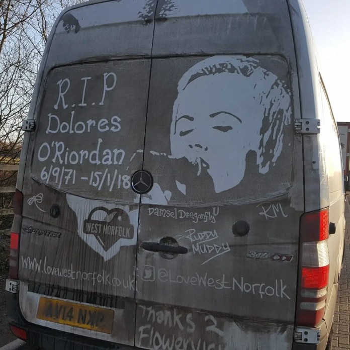 Grime art on the back of a van paying tribute to Dolores O'Riordan, the lead singer of The Cranberries, with the text 'R.I.P Dolores O'Riordan 6/9/71 - 15/1/18' and an image of her singing. The artwork includes additional tags such as 'West Norfolk,' '@LoveWestNorfolk,' and 'Thanks 2 Flowervision.' The artist has transformed the van’s dirty surface into a heartfelt memorial to the musician.