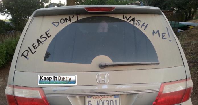 The back of a dusty Honda van with the phrase 'PLEASE DON'T WASH ME!' written in large letters across the rear window. A bumper sticker reads 'Keep It Dirty,' adding humor to the message and embracing the vehicle's dusty look as part of its character.