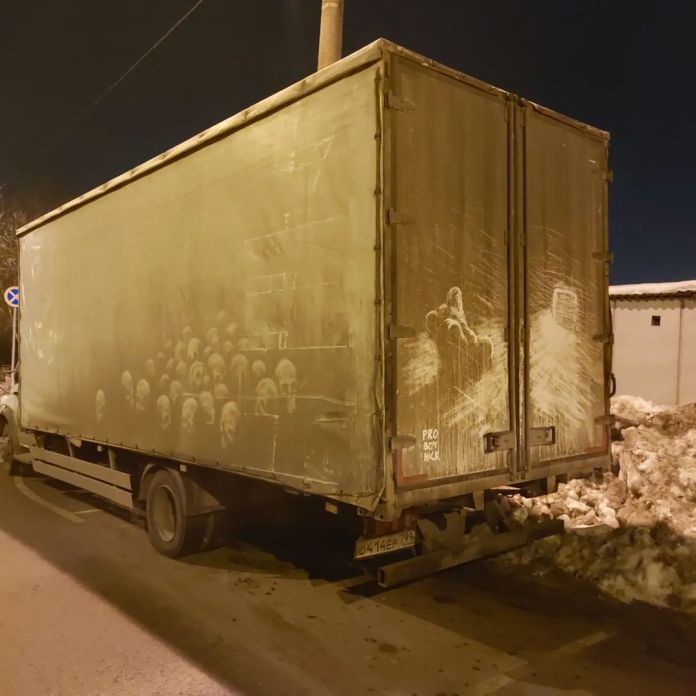 Grime art by Russian artist ProBoyNick on the side of a truck, featuring a haunting row of ghostly skulls etched into the dirt. The artwork extends across the vehicle, creating a somber and impactful display in the dim light of a nighttime street. This piece is a powerful anti-war statement in response to the conflict in Ukraine.