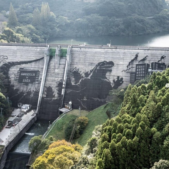 A breathtaking reverse graffiti artwork featuring Godzilla on the Iwayagawachi Dam in Saga Prefecture, Japan. The massive design depicts Godzilla towering over a cityscape, with intricate details of buildings and trees around him. The artwork was created as part of the "Godzilla in Saga Dam Art Project Powered by Kärcher," using high-pressure cleaning techniques to remove dirt and reveal the iconic monster's silhouette on the dam’s concrete surface. Surrounded by lush greenery and water, the dam serves as a striking canvas for this monumental tribute to Godzilla's legacy.