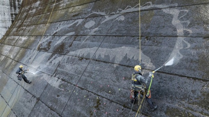 Two climbers from Kärcher creating a massive reverse graffiti artwork of Godzilla on the Iwayagawachi Dam in Saga Prefecture, Japan. Equipped with high-pressure cleaning tools, the climbers are suspended by ropes as they carefully remove dirt and moss to reveal the intricate design of Godzilla’s iconic silhouette. The process highlights the innovative technique of using cleaning as an artistic method.