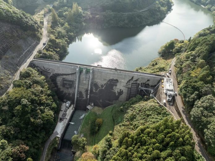 An aerial view of the Iwayagawachi Dam in Saga Prefecture, Japan, showcasing a massive reverse graffiti artwork of Godzilla. The iconic monster is depicted in a dramatic cityscape, with intricate details of buildings and surrounding greenery. The calm reservoir above the dam and lush forested hills add to the striking contrast of the dark artwork on the concrete structure. This monumental creation was part of the "Godzilla in Saga Dam Art Project Powered by Kärcher," blending nature, art, and technology in an unforgettable display.