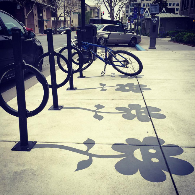 Public art installation by Damon Belanger in Redwood City, California, featuring creatively painted "fake shadows" beneath a row of circular bike racks. The black painted shadows transform the standard metal poles into whimsical flower stems and blooms, making the structures appear as if they're casting floral shadows on the sidewalk in bright daylight.
